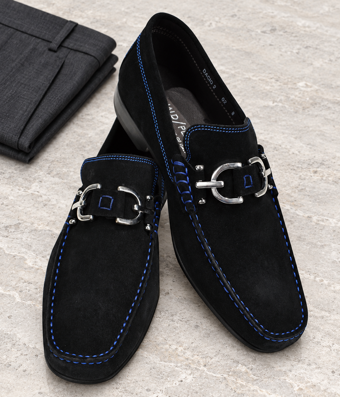 Black Dacio suede loafers with blue stitching and silver-tone hardware on pale stone beside folded charcoal trousers.