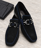 Black Dacio suede loafers with blue stitching and silver-tone hardware on pale stone beside folded charcoal trousers.