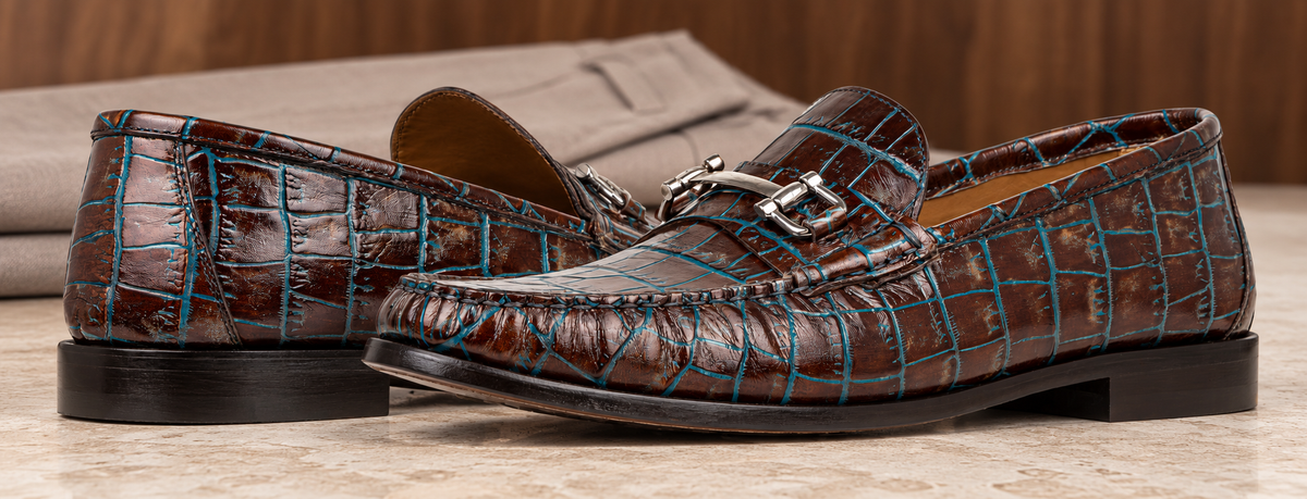 Donald Pliner Evan94 chocolate loafers with turquoise pattern accents on stone beside folded taupe trousers.
